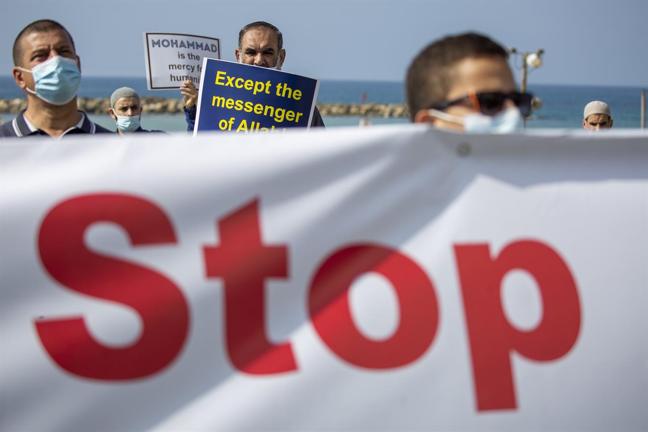 Israeli Arab protesters wearing face masks due to the coronavirus pandemic hold signs during a protest against French President Emmanuel Macron and the publishing of caricatures of the Muslim Prophet Muhammad they deem blasphemous, in front of the French embassy in Tel Aviv, Israel, Tuesday, Oct. 27, 2020. (AP Photo/Ariel Schalit)
