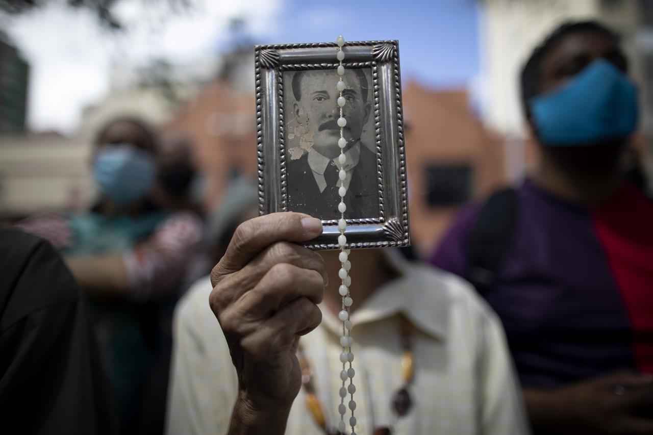 A devotee holds up a framed image of the late Dr. Jose Gregorio Hernandez outside La Candelaria church where his remains are interred, in Caracas, Venezuela, Monday, Oct. 26, 2020. The remains of Hernandez, popularly known as the "doctor of the poor", were exhumed on Monday in a closed ceremony in a church in downtown Caracas as part of the Vatican's demands for the beatification of the first Venezuelan layman. (AP Photo/Ariana Cubillos)
