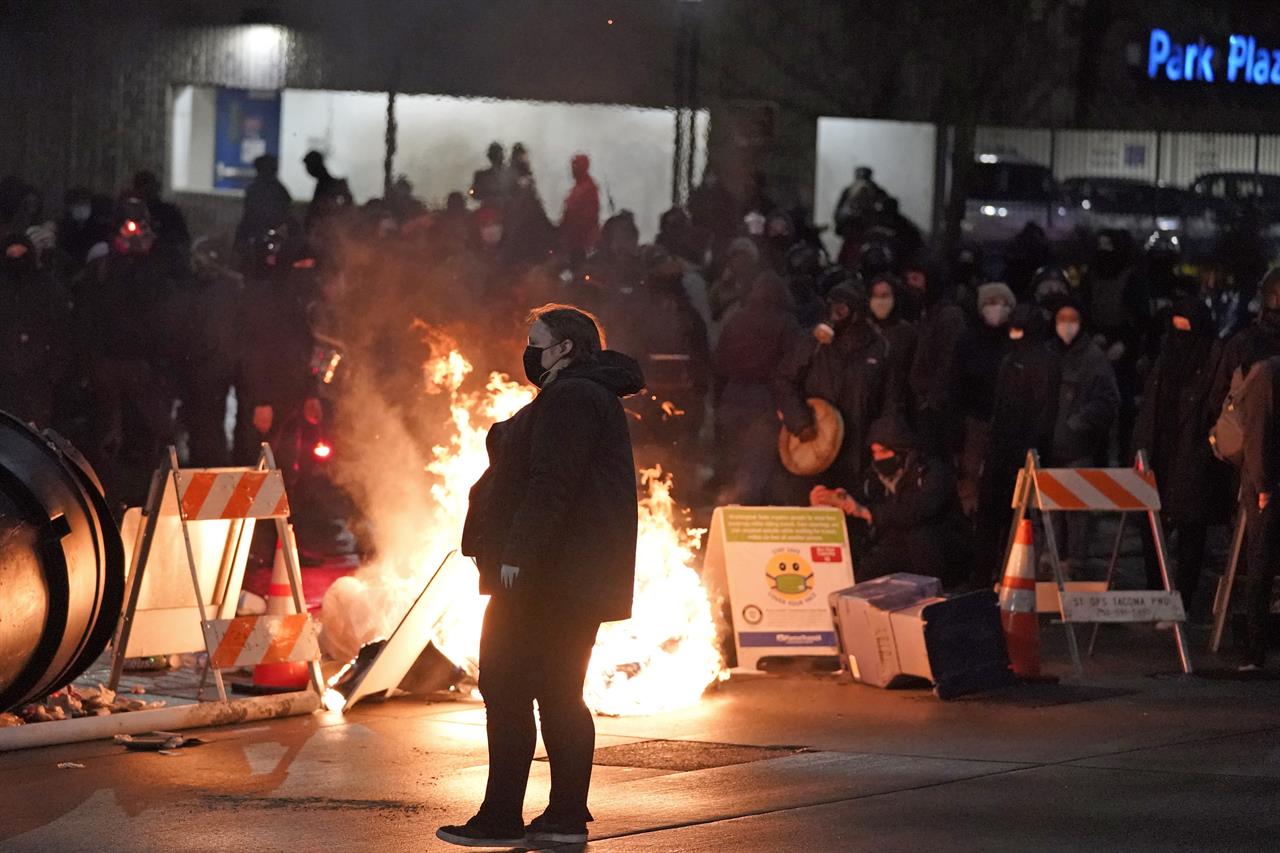 A protester walks past burning trash during a protest against police brutality, late Sunday, Jan. 24, 2021, in downtown Tacoma, Wash., south of Seattle. The protest came a day after at least two people were injured when a Tacoma Police officer responding to a report of a street race drove his car through a crowd of pedestrians that had gathered around him. Several people were knocked to the ground and at least one person was run over. (AP Photo/Ted S. Warren)